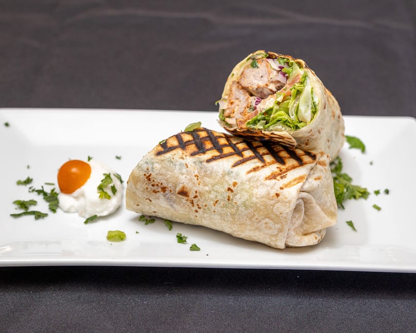 Grilled chicken wrap, cut in half, with lettuce, red onion, and a side of white sauce with a cherry tomato.