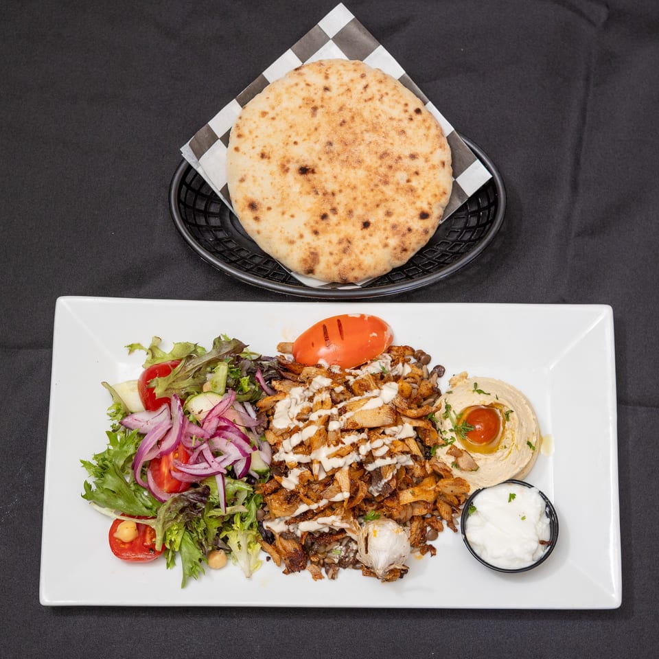 Chicken shawarma plate with salad, hummus, garlic sauce, and pita bread on a black background.