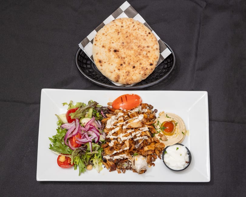 Chicken shawarma plate with salad, hummus, garlic sauce, and pita bread on a black background.