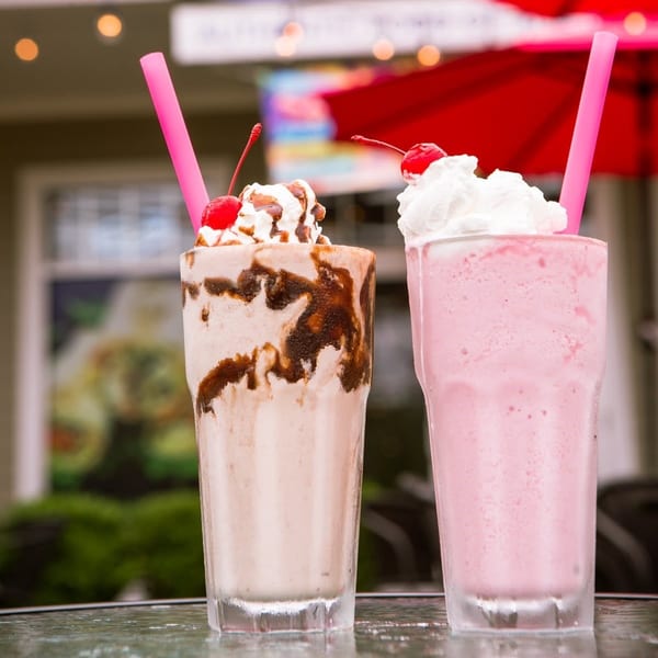 Two milkshakes, one chocolate and one strawberry, topped with whipped cream and cherries on a table.