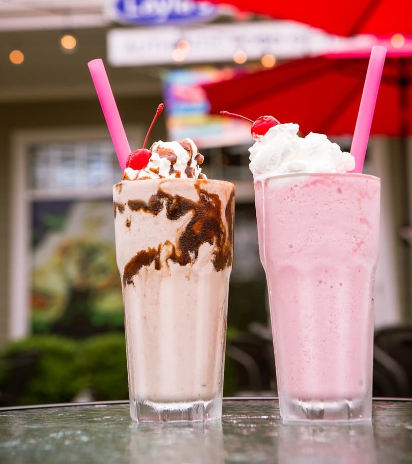 Two milkshakes, one chocolate and one strawberry, topped with whipped cream and cherries on a table.