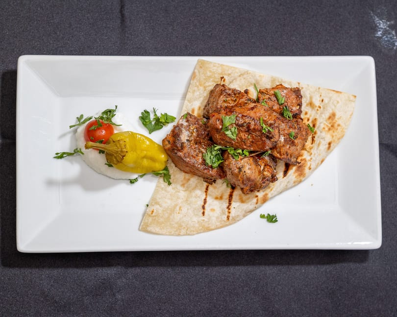 Grilled meat pieces with parsley on flatbread, served with a cherry tomato, pepper, and white sauce.