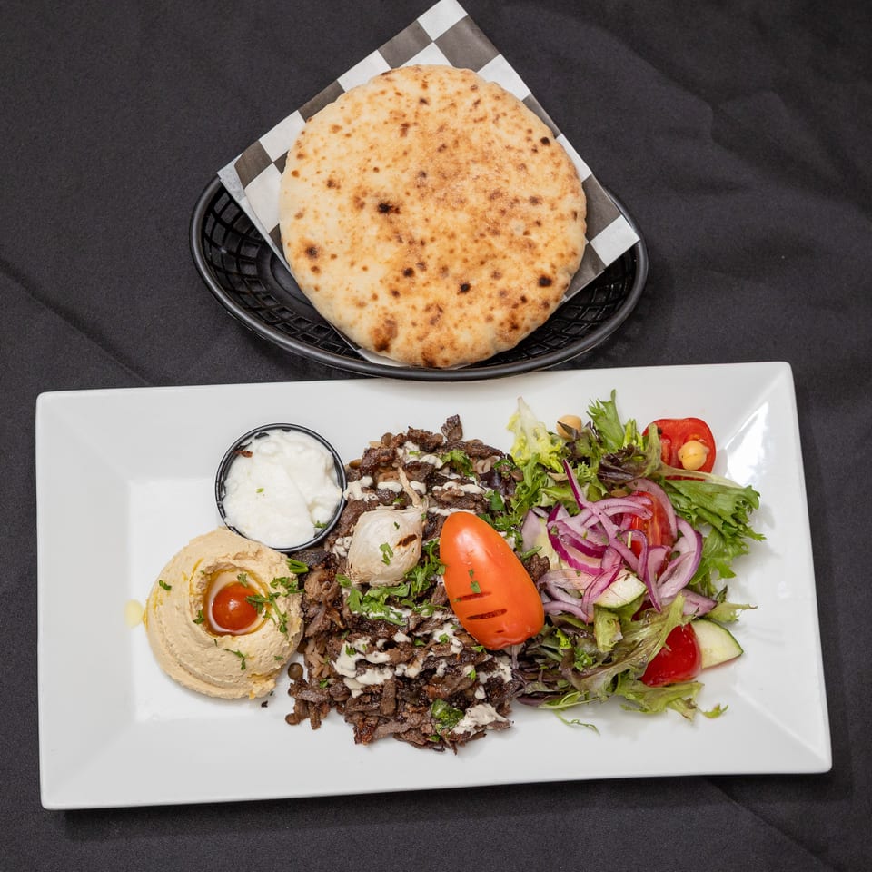 Shawarma plate with meat, hummus, salad, garlic sauce, and pita bread on a black background.