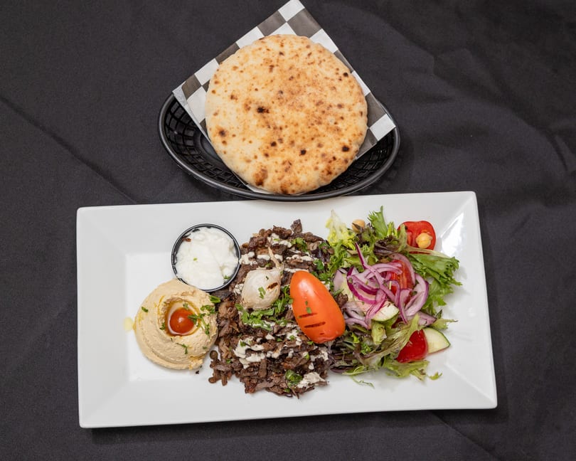 Shawarma plate with meat, hummus, salad, garlic sauce, and pita bread on a black background.