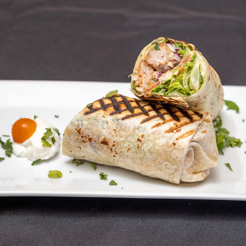 Grilled chicken wrap, cut in half, with lettuce, red onion, and a side of white sauce with a cherry tomato.