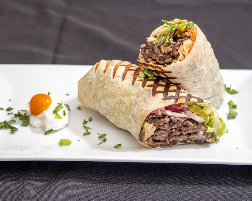 Two grilled shawarma wraps filled with meat, lettuce, and onions, served with a side of sauce.