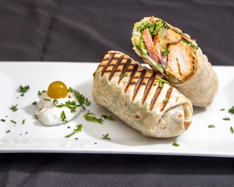 Two grilled chicken wraps with fresh vegetables, one cut open, served with a dollop of sauce.