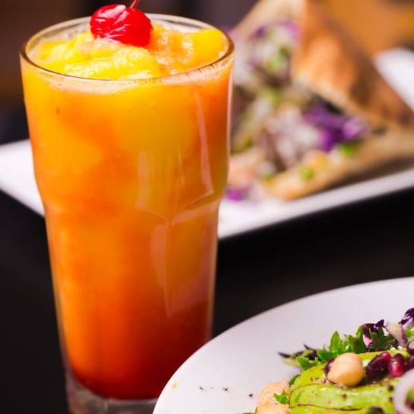 A vibrant orange and yellow drink with a cherry, next to a fresh salad and a sandwich.
