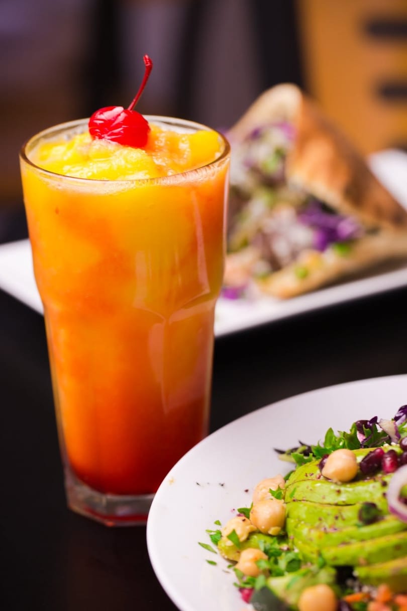 A vibrant orange and yellow drink with a cherry, next to a fresh salad and a sandwich.