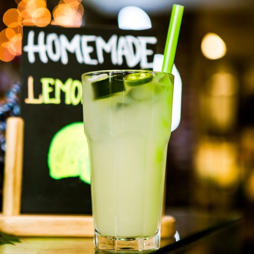 Homemade lemonade with cucumber slices and a green straw, next to a chalkboard sign.
