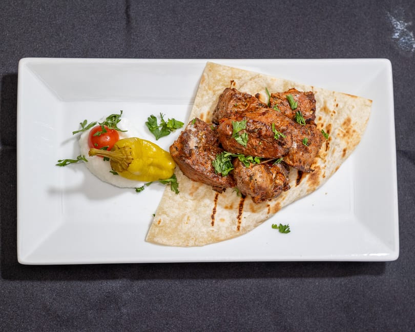 Grilled meat pieces with parsley on flatbread, served with a cherry tomato, pepper, and white sauce.