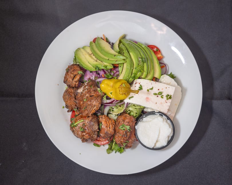 Grilled meat, avocado, feta cheese, and salad with a side of white sauce on a white plate.