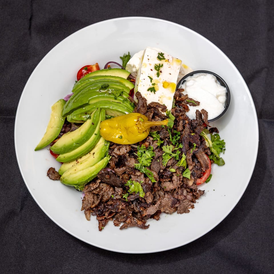 A white plate with a large serving of sliced meat, avocado, feta cheese, and a side of white sauce.