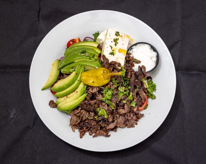A white plate with a large serving of sliced meat, avocado, feta cheese, and a side of white sauce.